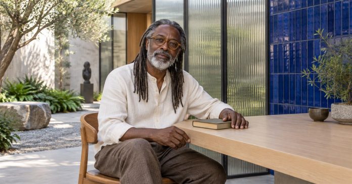 A quiet reflective moment in a cultural-centre courtyard Man with salt-and-pepper locs and amber glasses seated beside a closed book in a courtyard.